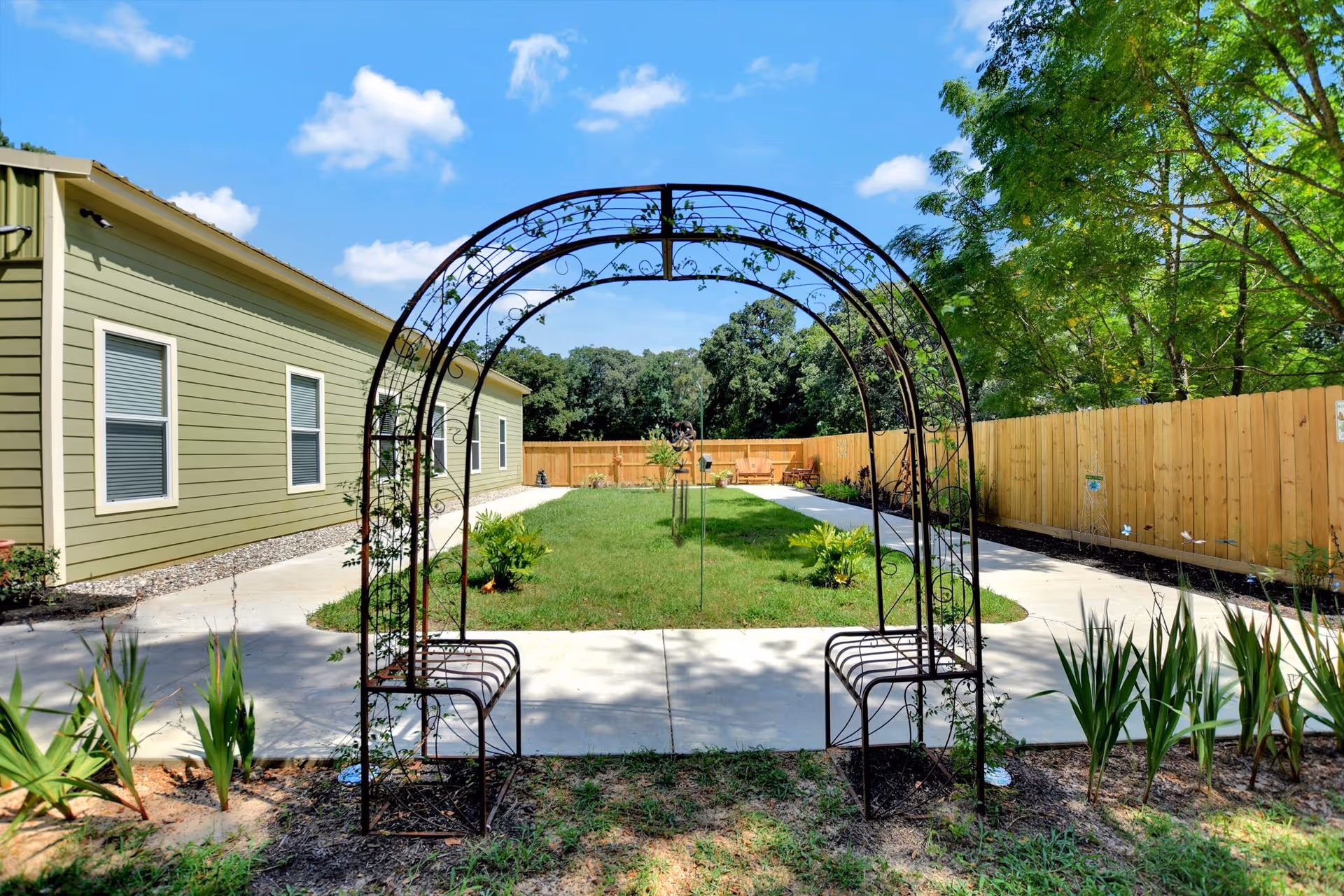 Wrought-iron garden arch and benches framing a grassy courtyard with a pathway, wooden fence, and a light green building under a blue sky.