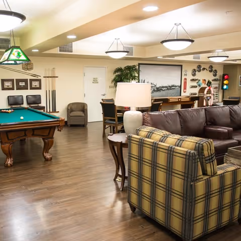 A spacious senior living common room with a pool table, seating area with sofas and armchairs, a bar, and wall decor.