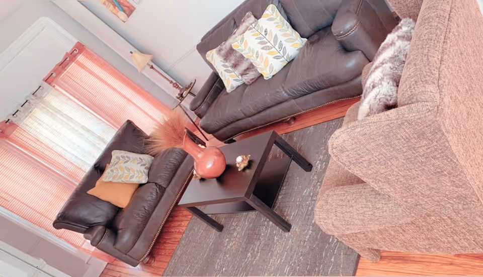 A cozy living room with two dark brown leather sofas and one light brown fabric armchair arranged around a black coffee table. The coffee table has a decorative vase with dried plants and a small ornament. The room has wooden floors, a gray area rug, pink curtains, and a floor lamp in the corner.