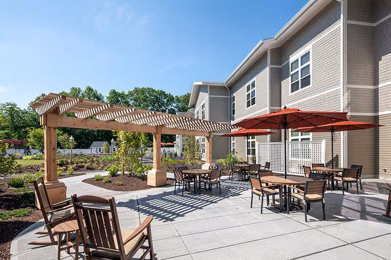 Outdoor patio with a wooden pergola, tables and chairs, and red umbrellas beside a multi-story building.