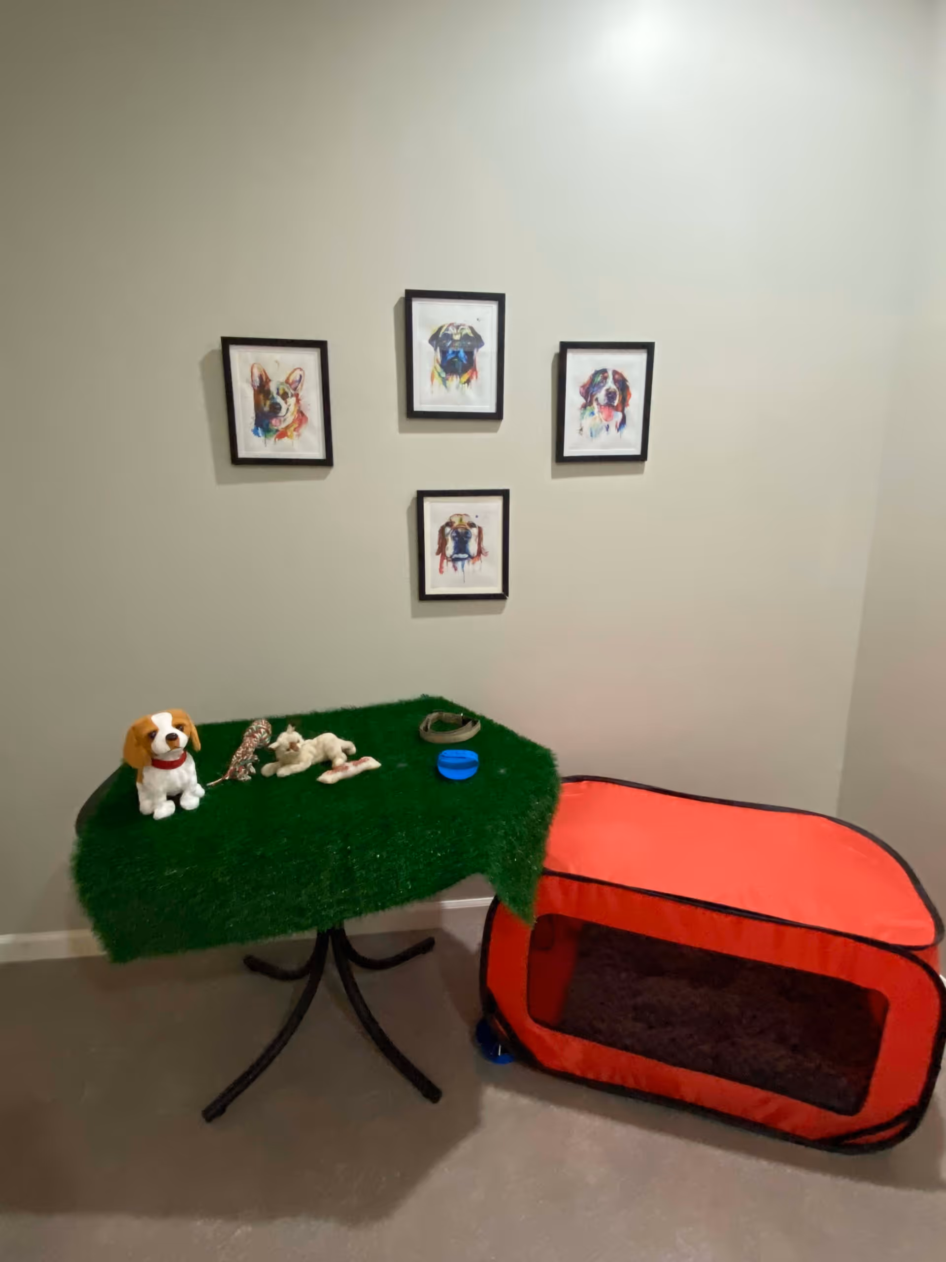 A small indoor corner with a table covered in green artificial grass holding dog toys and a small dog figurine. Next to the table is a red pet crate with a dark cushion inside. On the wall above are four framed colorful paintings of dogs.