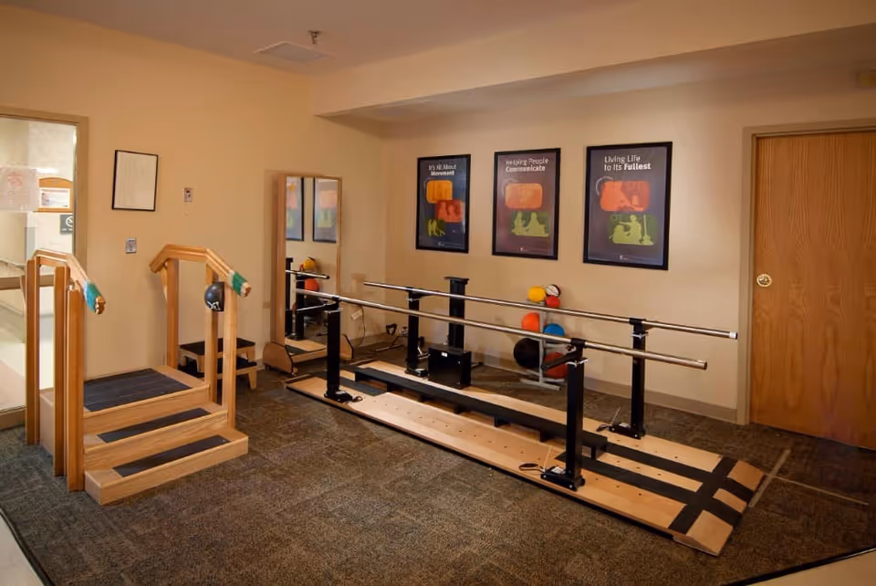 A rehabilitation or physical therapy room with parallel bars for walking exercises, a small wooden staircase with handrails, a large mirror, and colorful therapy balls stacked in the corner. Three motivational posters are displayed on the wall.