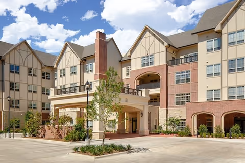 Multi-story senior living building with a covered porte-cochere entrance, brick and stucco facade, landscaped driveway and blue sky.