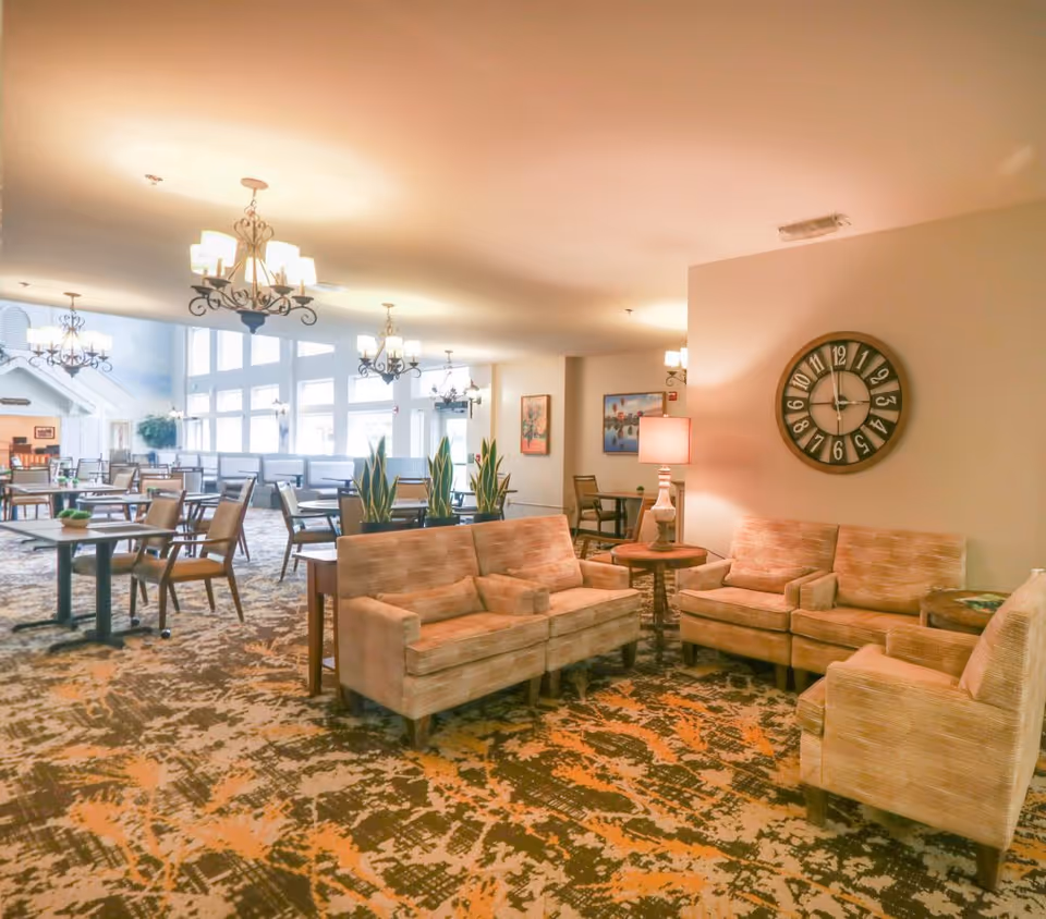 A spacious and well-lit common area in a senior living facility featuring beige upholstered sofas and armchairs arranged around wooden side tables with lamps. The room has patterned carpet flooring, several chandeliers hanging from the ceiling, large windows letting in natural light, and a large decorative wall clock. There are also dining tables and chairs in the background along with some potted plants and framed artwork on the walls.
