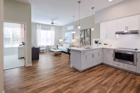Bright and modern open-concept living space featuring a kitchen with white cabinetry, a large island with pendant lighting, and stainless steel appliances. Adjacent to the kitchen is a living room area with a sofa, coffee table, and large windows allowing natural light to fill the room. A separate room with carpet flooring is visible through an open doorway.