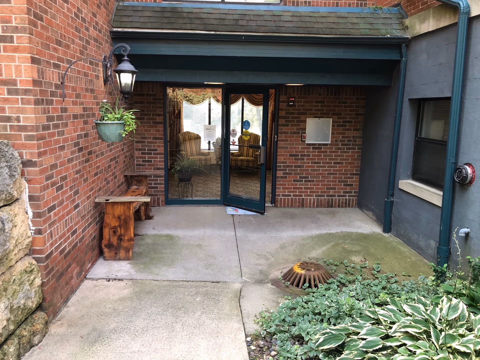 Covered brick entrance with glass double doors opening to a seating area, a wooden bench, hanging planter, and a drain in the foreground.