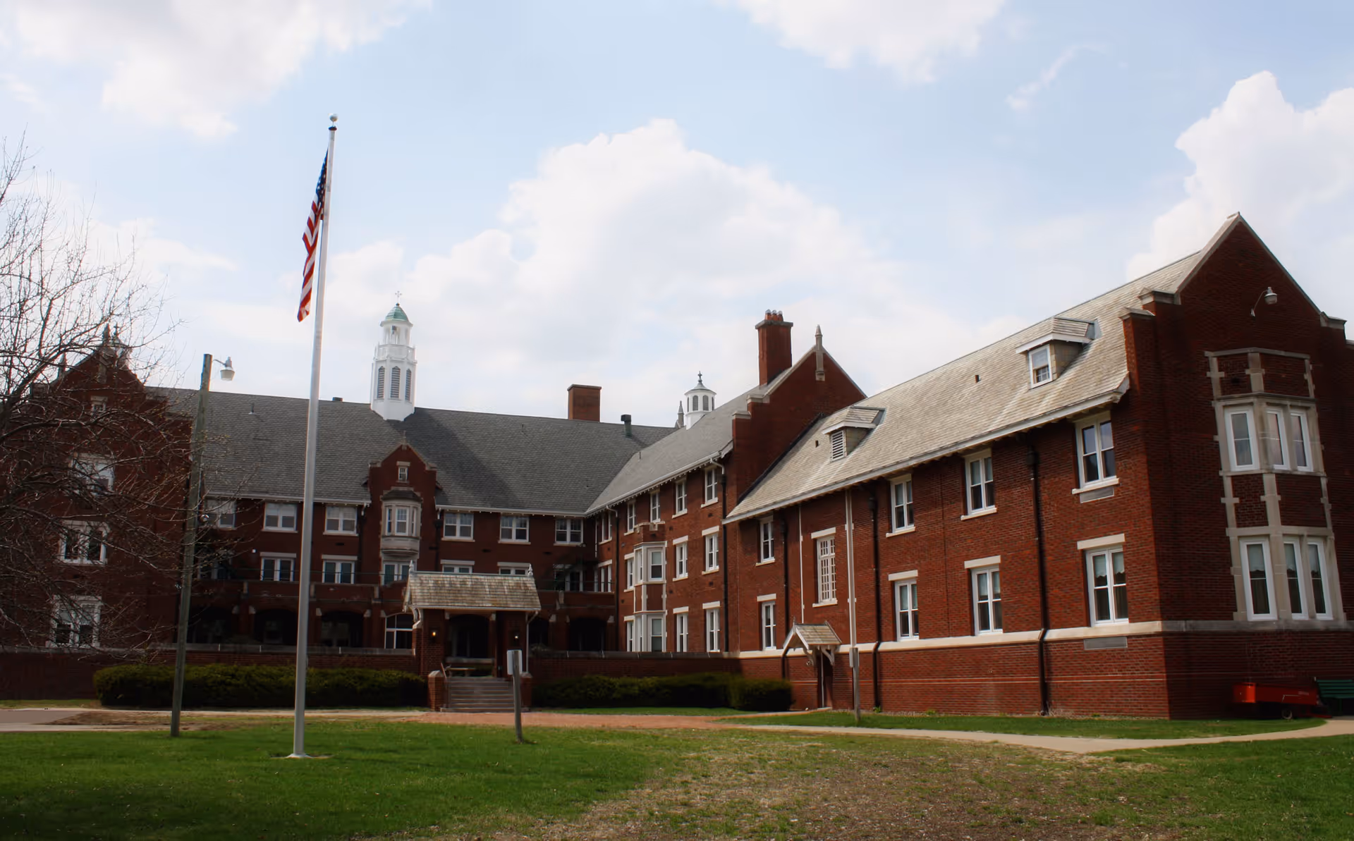 Exterior view of a large, multi-story brick building with white-trimmed windows and a gray roof. There is an American flag on a flagpole in front of the building, along with some bushes and a grassy area. The sky is partly cloudy.