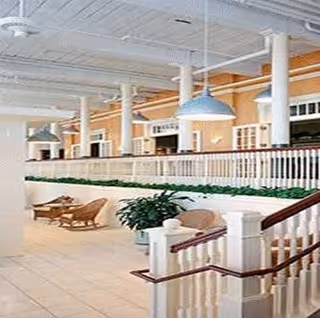 Bright interior common area with white columns and railings, pendant lights, potted plants, and seating along a tiled floor.