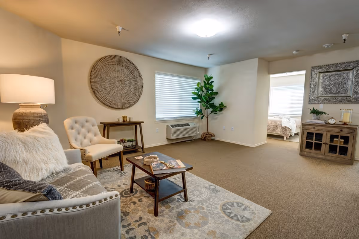 A cozy living room area with a beige sofa adorned with pillows and a throw blanket, a cushioned chair, a wooden coffee table on a patterned rug, a side table with a lamp, a large decorative wall piece, a potted plant near a window with blinds, and an air conditioning unit below the window. In the background, there is an open doorway leading to a bedroom with a bed and a wooden cabinet with decorative items.