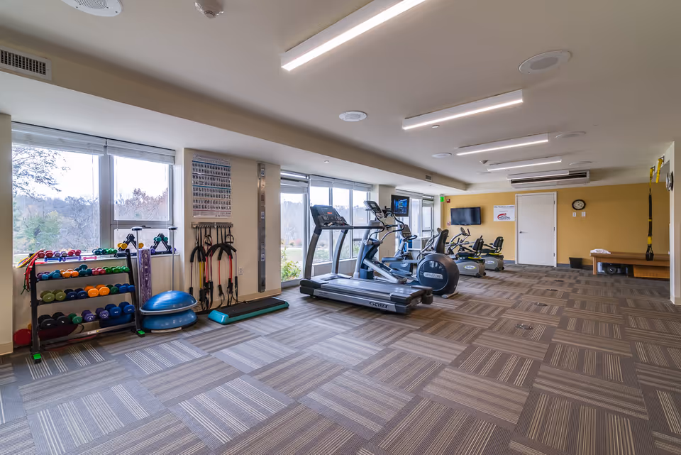 A spacious fitness room with large windows letting in natural light. The room contains various exercise equipment including treadmills, stationary bikes, dumbbells, resistance bands, balance trainers, and a wall-mounted TV. The floor is carpeted with a patterned design, and the walls are painted in neutral tones with one yellow accent wall.