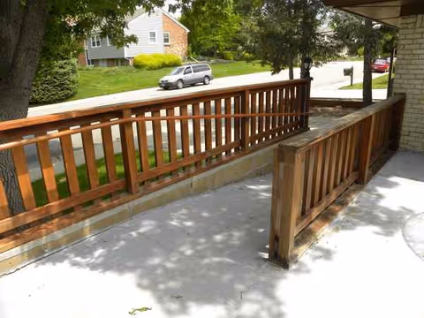 Wooden railing along a concrete outdoor porch with trees and a street view.
