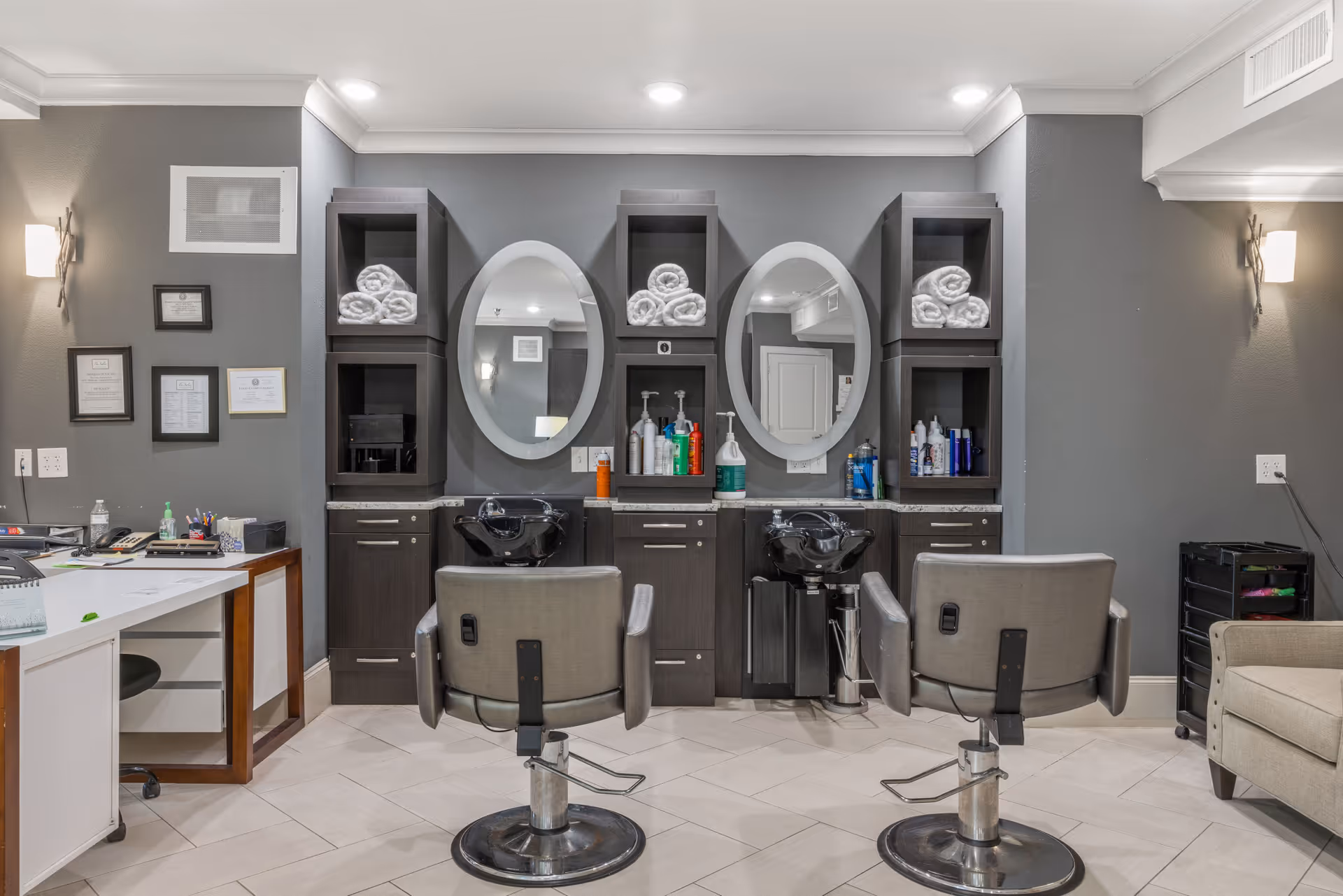 Interior view of a salon area in a senior living facility featuring two salon chairs facing black wash basins with oval mirrors above. The walls are painted gray, and there are shelves with rolled white towels and various hair care products. To the left, there is a desk with office supplies and framed certificates on the wall. The floor is tiled in a light color.