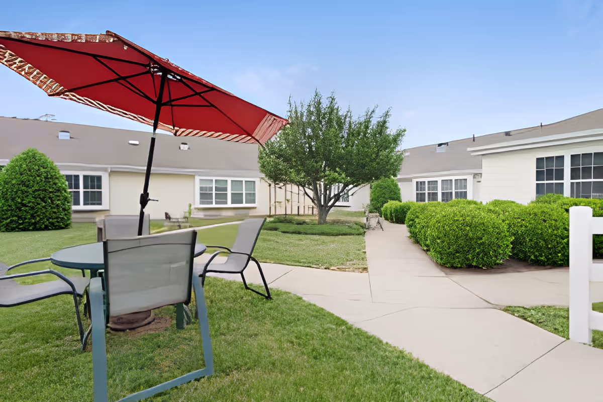 Outdoor courtyard with a round table, chairs and a red patio umbrella, walkways, trimmed bushes and a one-story building in the background.