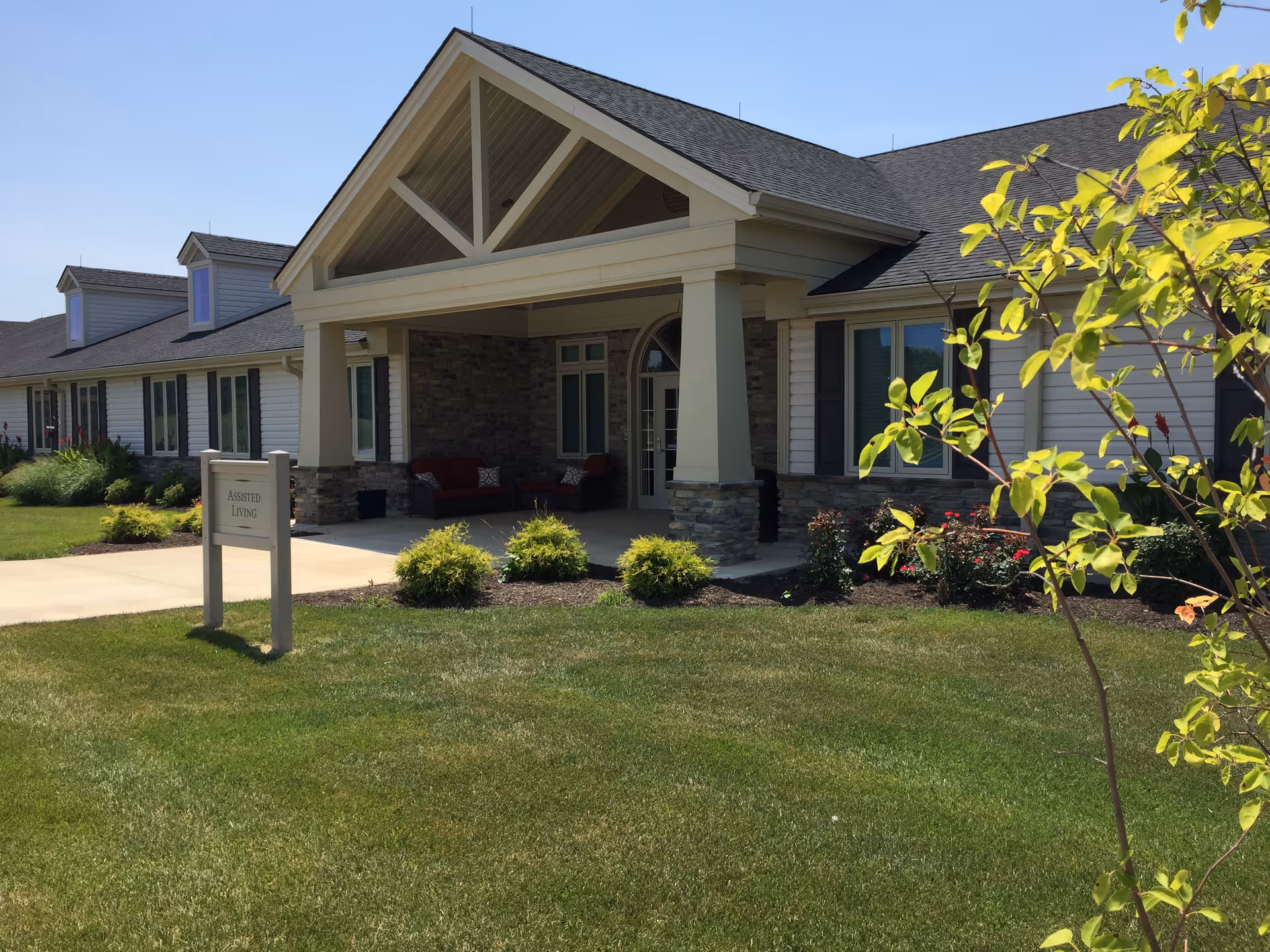 Exterior view of the Otterbein Cridersville SeniorLife Community assisted living building with a covered entrance, stone and siding facade, a well-maintained lawn, shrubs, and a small tree in the foreground under a clear blue sky.