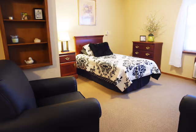 A cozy bedroom in Wheeler Terrace featuring a single bed with a black and white patterned bedspread and black pillows. The room includes a wooden nightstand with a lamp, a wooden dresser with decorative items and a framed picture on the wall. There are two black armchairs and a window with white curtains allowing natural light into the room.