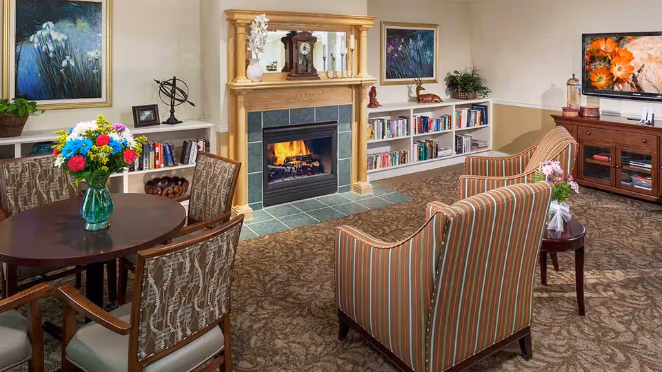 A cozy living room area with a lit fireplace framed by a wooden mantel. There are two striped armchairs facing the fireplace, a round table with a vase of colorful flowers, and a wooden cabinet with a flat-screen TV displaying an image of orange flowers. Bookshelves filled with books and decorative items line the walls, and framed artwork hangs above the shelves. The room has patterned carpet and neutral-colored walls.