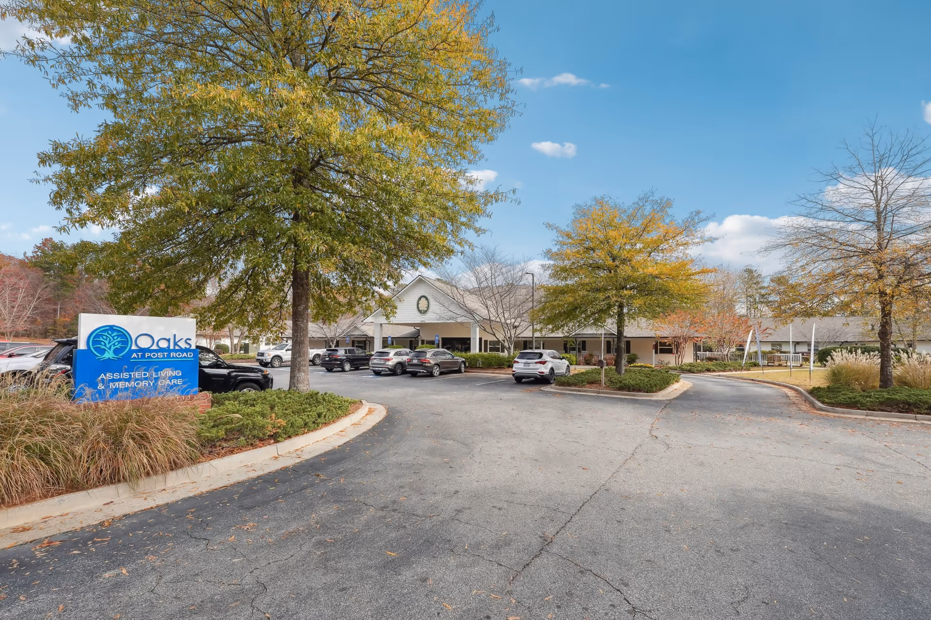 Exterior view of Oaks at Post Road assisted living and memory care facility with a parking lot, several cars, trees with autumn foliage, and a clear blue sky.