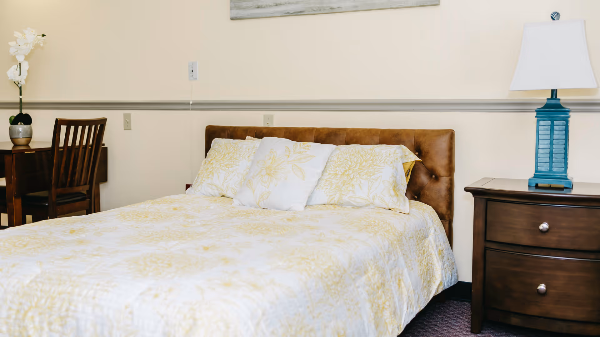 A bedroom with a bed featuring a brown tufted headboard, yellow and white floral bedding, and three pillows. To the right of the bed is a wooden nightstand with two drawers and a blue lamp with a white lampshade. To the left is a wooden chair and desk with a potted white orchid on top. The walls are light-colored with a chair rail molding.