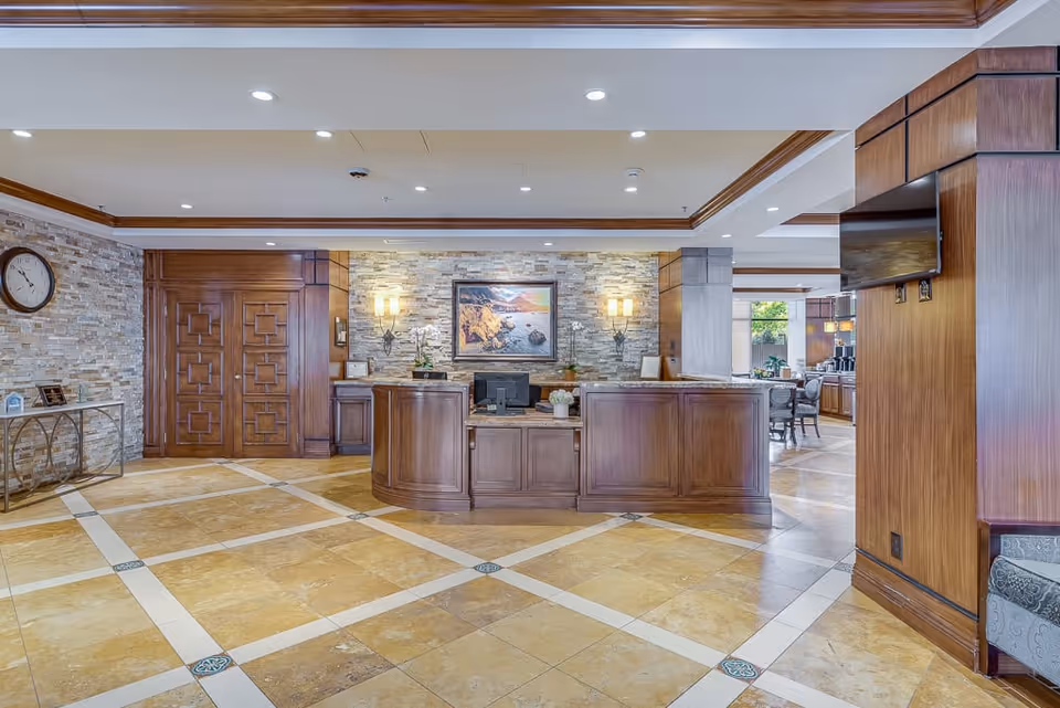 Reception area of The Park Lane facility featuring a wooden front desk with a computer, stone accent wall with a framed painting and two wall sconces, tiled floor with a geometric pattern, wooden double doors on the left, a clock on the wall, and a television mounted on a wooden column on the right.