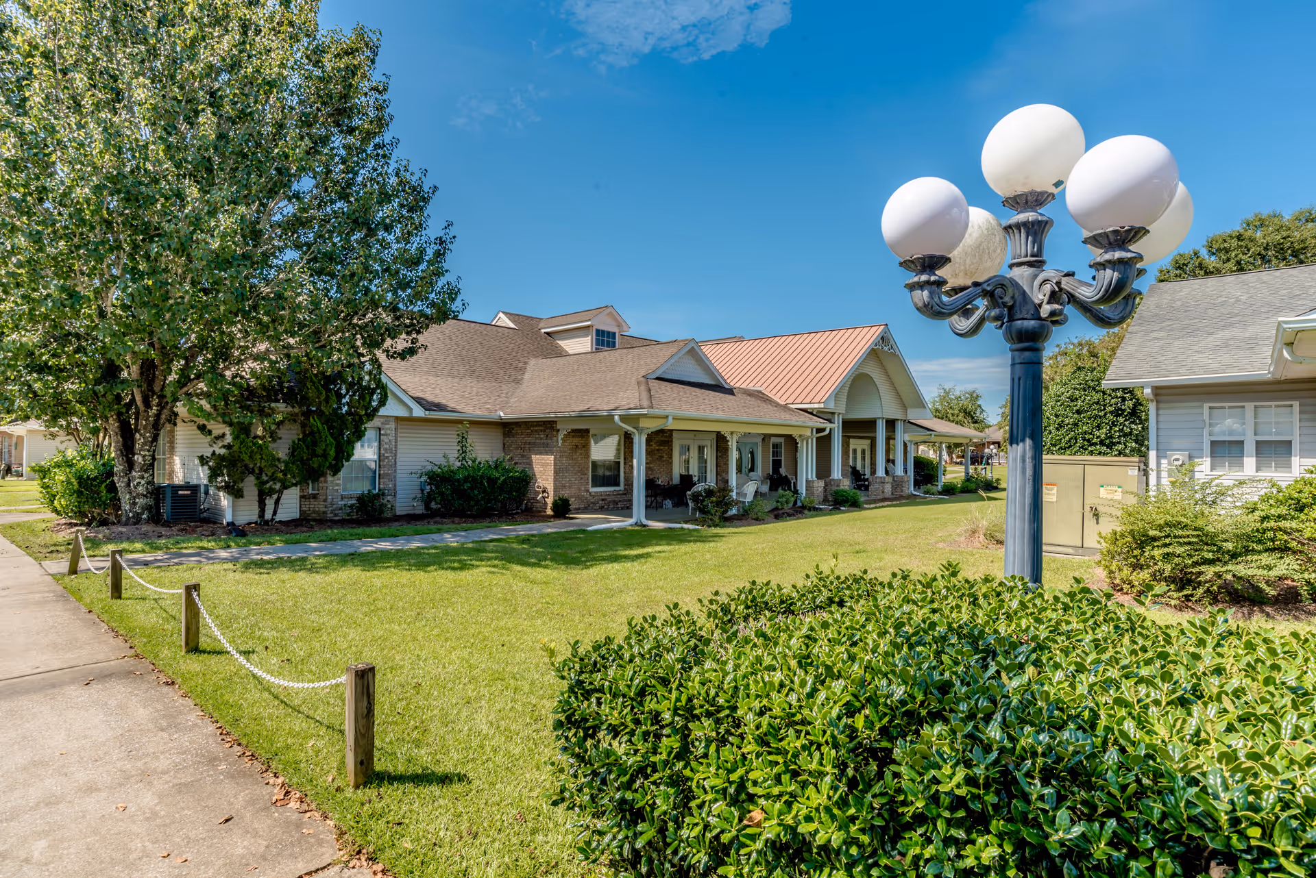 Exterior view of a senior living facility building with a well-maintained lawn, bushes, a large tree, and a decorative street lamp with five globe lights under a clear blue sky.