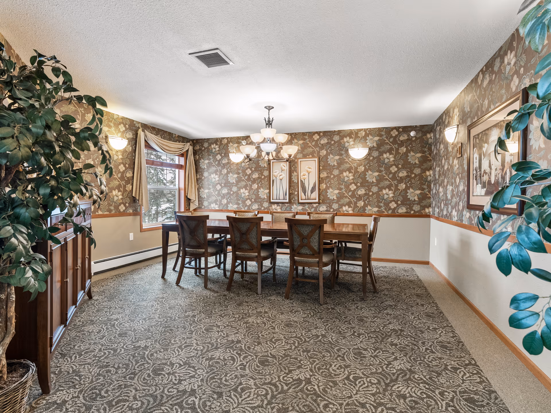 Formal dining room with a wooden table and chairs, floral wallpaper, chandelier, and potted plants.