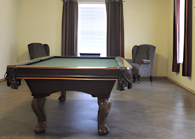 A pool table centered in a sparsely furnished room with two armchairs and curtained windows.