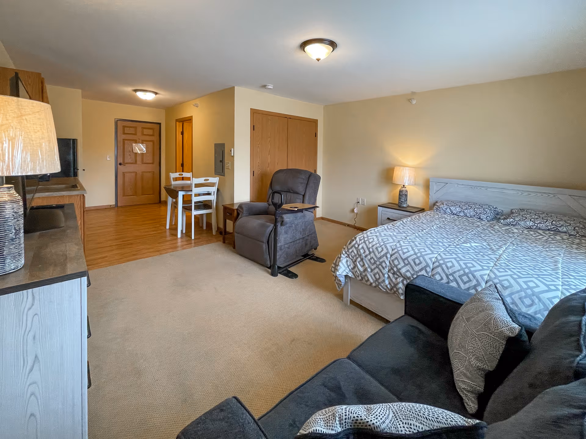 A cozy senior living studio apartment featuring a bed with patterned bedding, a reclining chair with an attached tray, a small dining table with two chairs, a sofa with cushions, a TV on a stand, and warm lighting from two lamps. The room has beige walls and a mix of carpet and wood flooring.