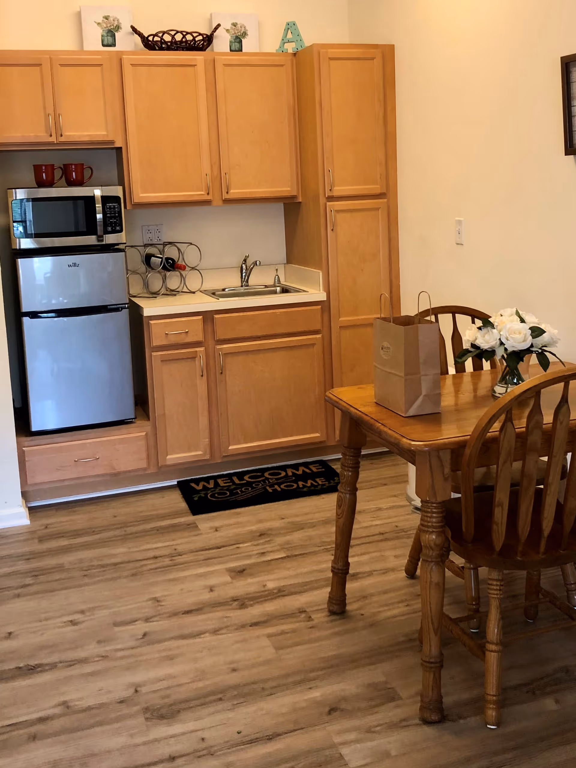 A small kitchen area with light wood cabinets, a stainless steel mini refrigerator, a microwave, a sink, and a wine rack on the counter. To the right, there is a wooden dining table with two chairs, a paper bag, and a vase with white flowers. The floor is wood, and there is a black welcome mat in front of the kitchen area.