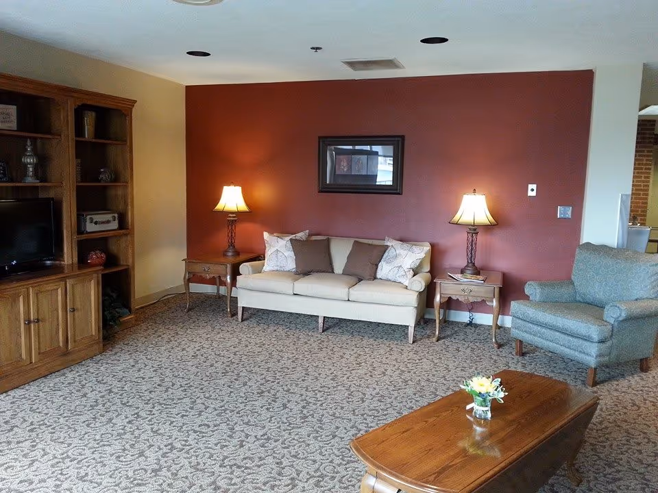 A cozy living room with a beige sofa adorned with pillows, flanked by two wooden side tables each holding a lamp. To the right is a patterned armchair, and to the left is a wooden entertainment center with a TV and decorative items. A wooden coffee table with a small flower arrangement sits in the foreground. The back wall is painted a deep red with a framed picture hanging above the sofa.