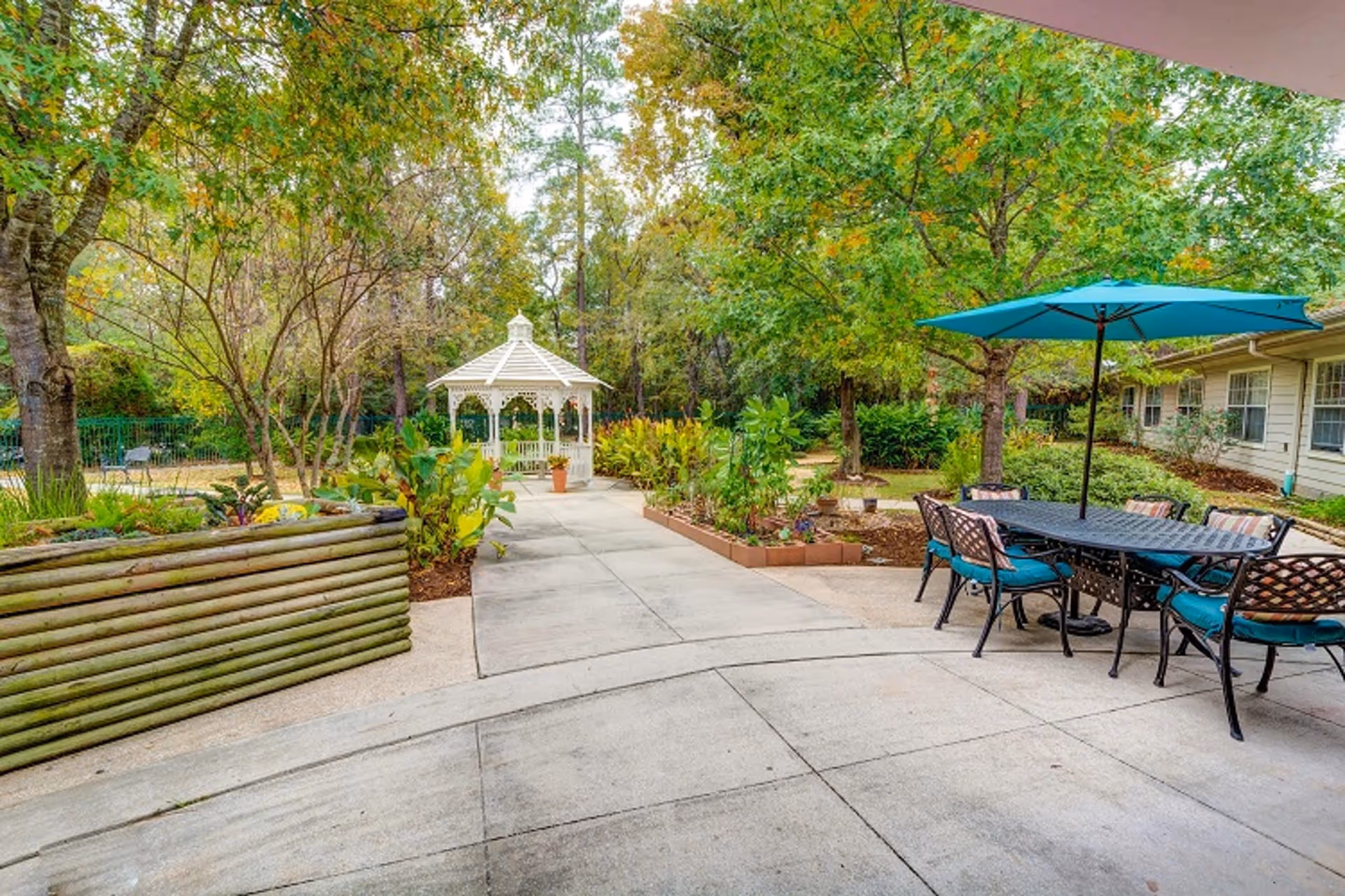 Outdoor patio area with a black metal table and six chairs with teal cushions under a large teal umbrella. A concrete pathway leads to a white gazebo surrounded by green trees and plants. Raised garden beds with various plants are visible along the pathway.