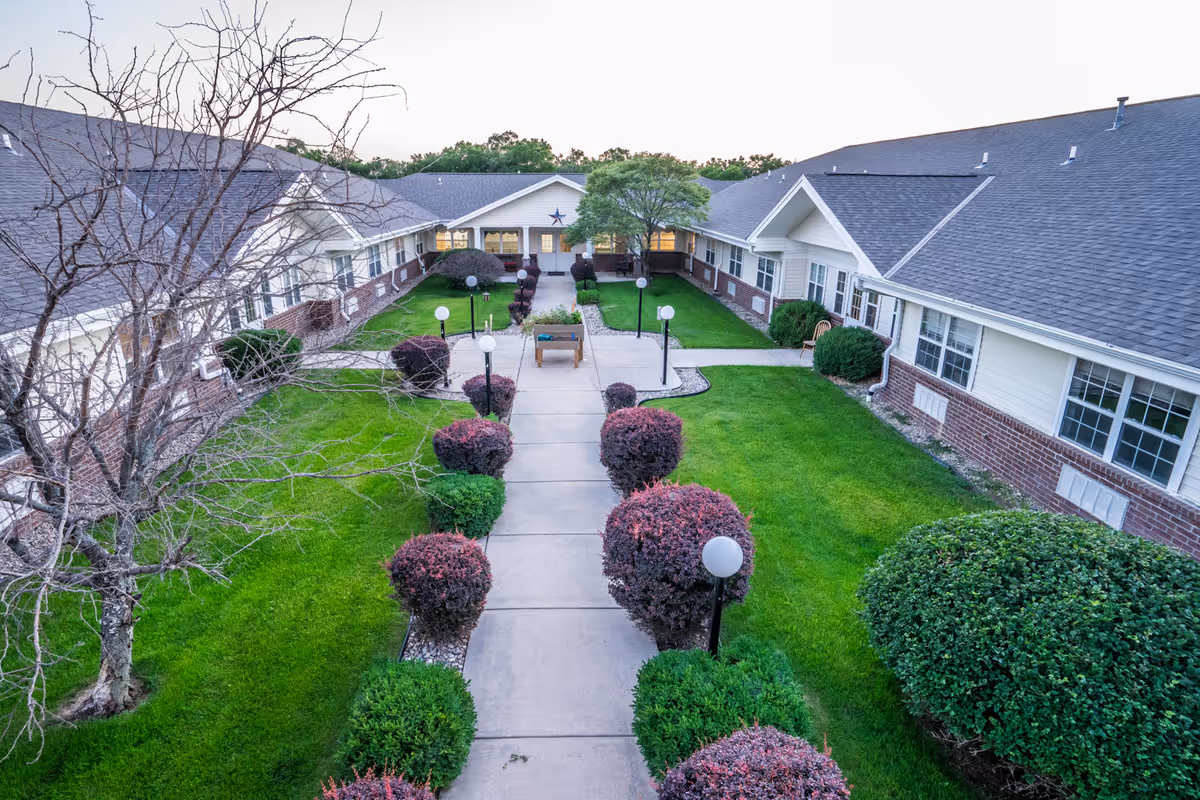 View of a courtyard at Saunders House featuring a paved walkway lined with trimmed bushes and lamp posts, surrounded by green lawns and a two-wing building with white siding and brick accents.