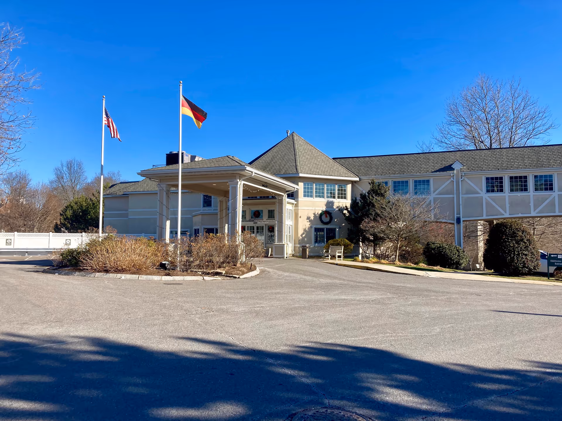 Exterior view of the German Centre Home building with two flagpoles displaying the American and German flags. The building has a covered entrance, beige walls, and a connecting walkway. There are bushes and trees around the entrance, and the sky is clear and blue.
