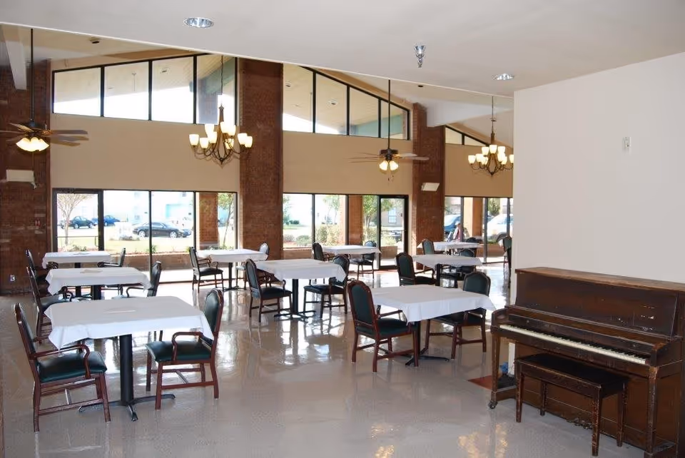 A spacious dining room with multiple tables covered with white tablecloths and surrounded by dark wooden chairs with green cushions. The room features large windows letting in natural light, brick pillars, ceiling fans, chandeliers, and an upright piano with a matching bench against a white wall.
