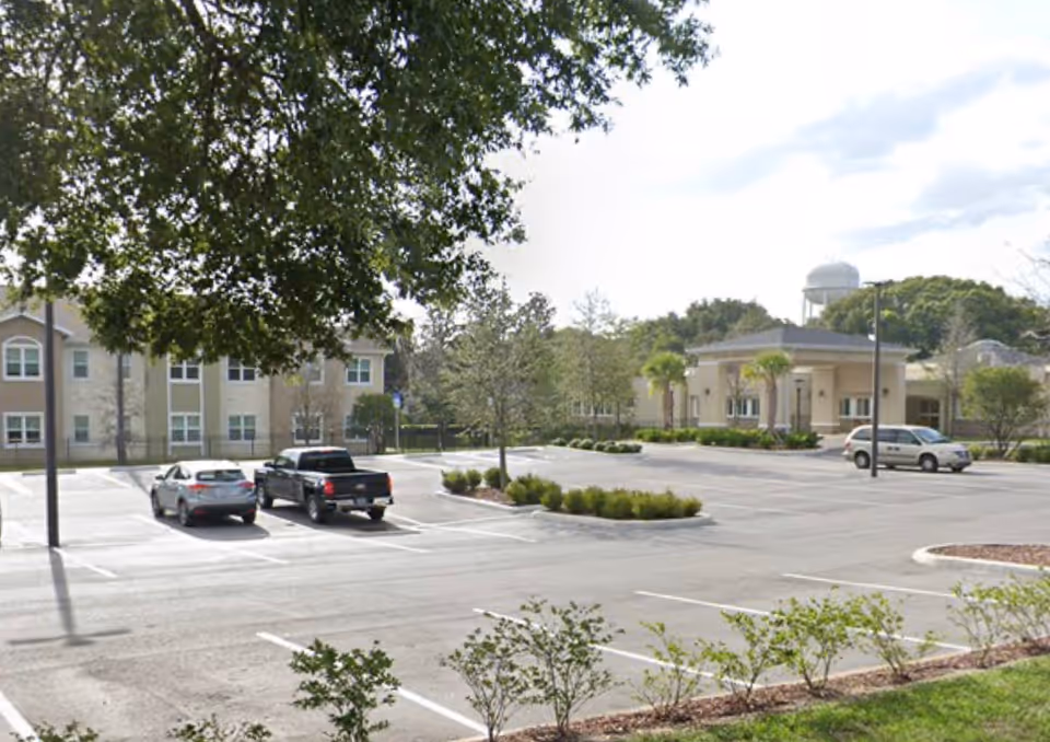 A mostly empty parking lot with a few cars in front of a beige senior living building entrance, surrounded by trees and landscaping.