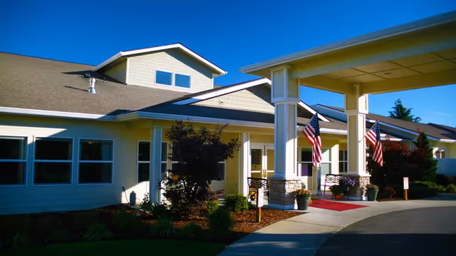Front entrance and covered drop-off of a single-story senior living building with American flags and landscaped beds.