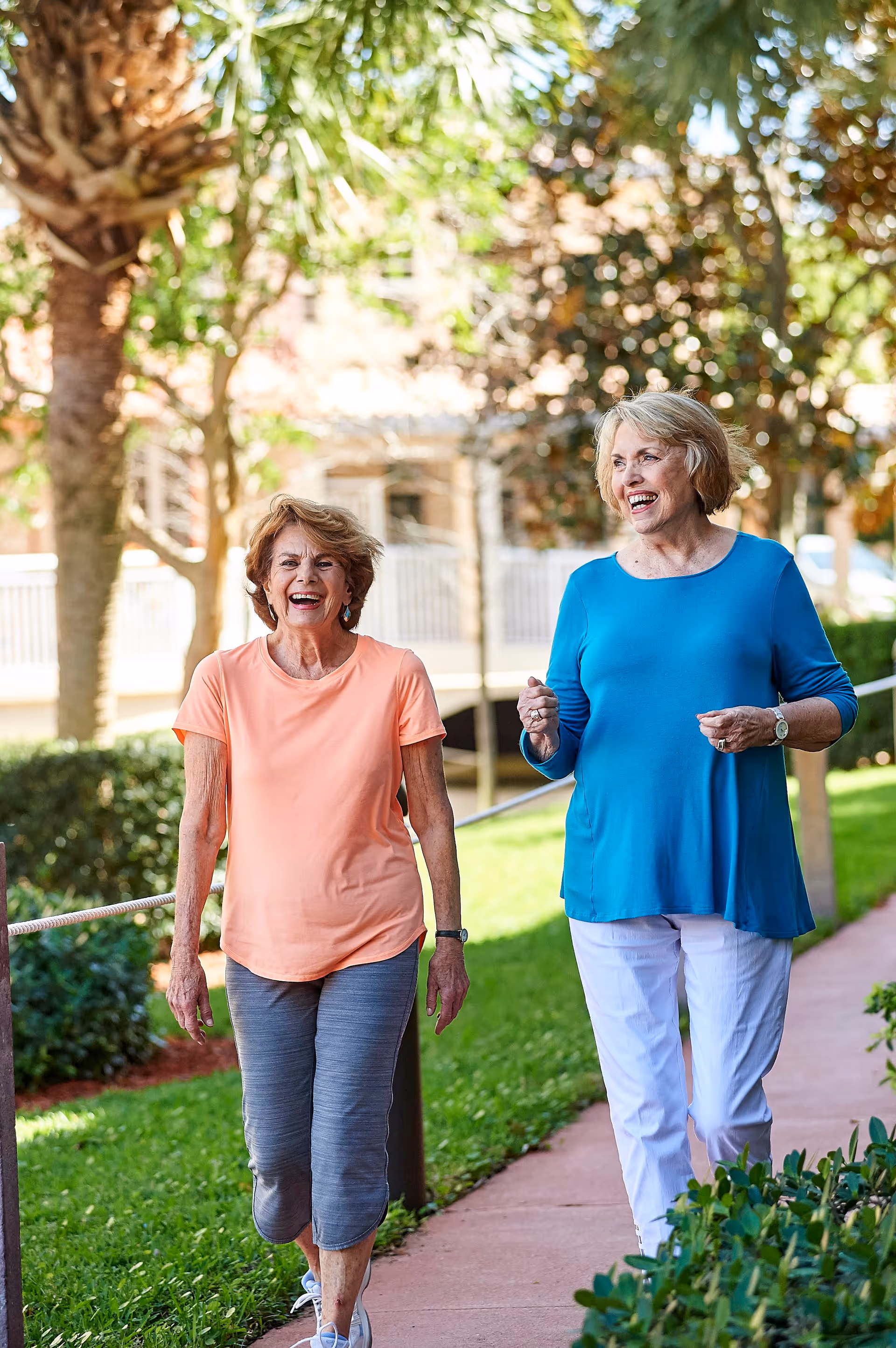 Two elderly women walking and smiling along a paved pathway in a sunny outdoor garden area with trees and bushes around them.