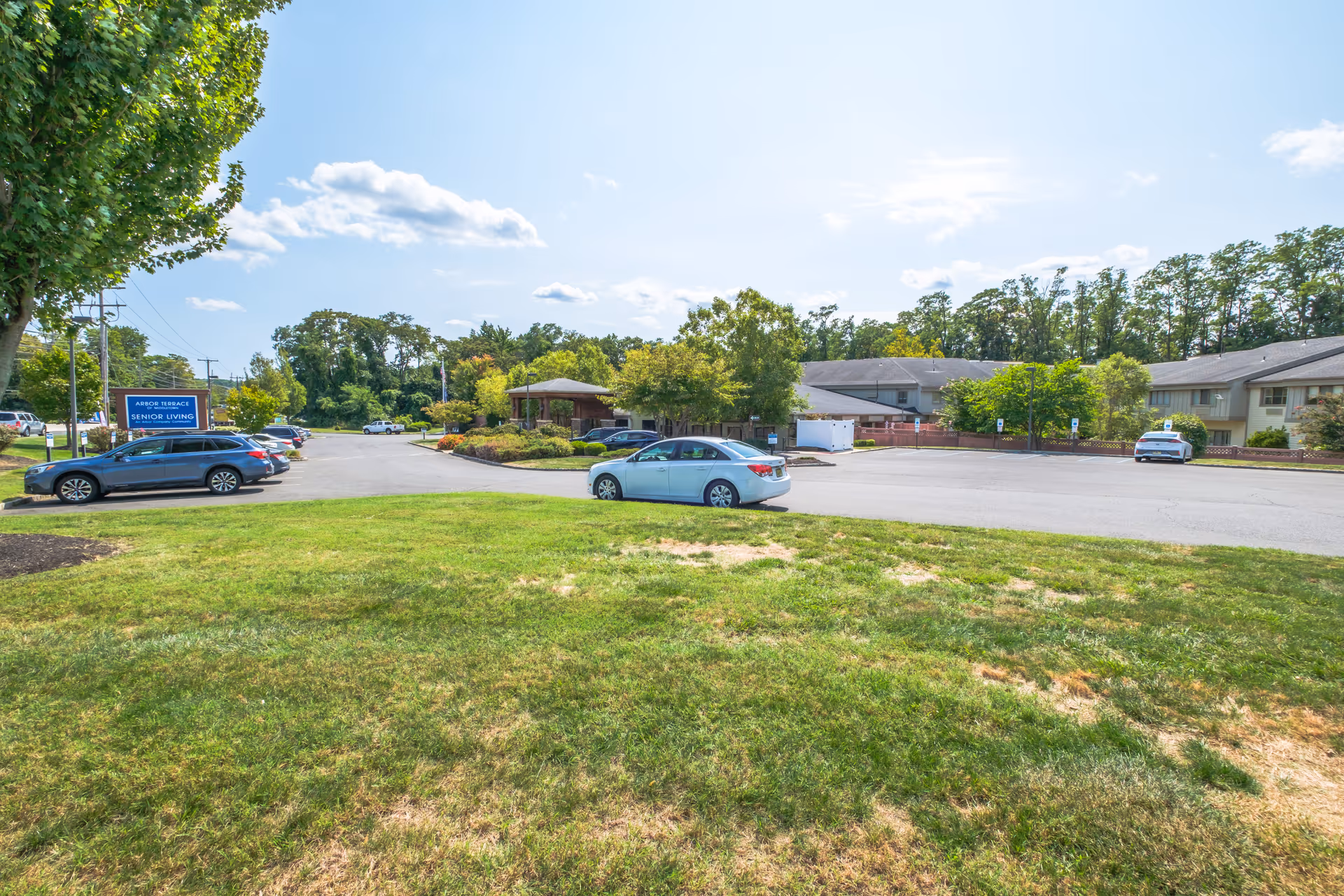 Parking lot area of Arbor Terrace Middletown senior living facility with several parked cars, green grass in the foreground, trees, and a clear blue sky with some clouds.