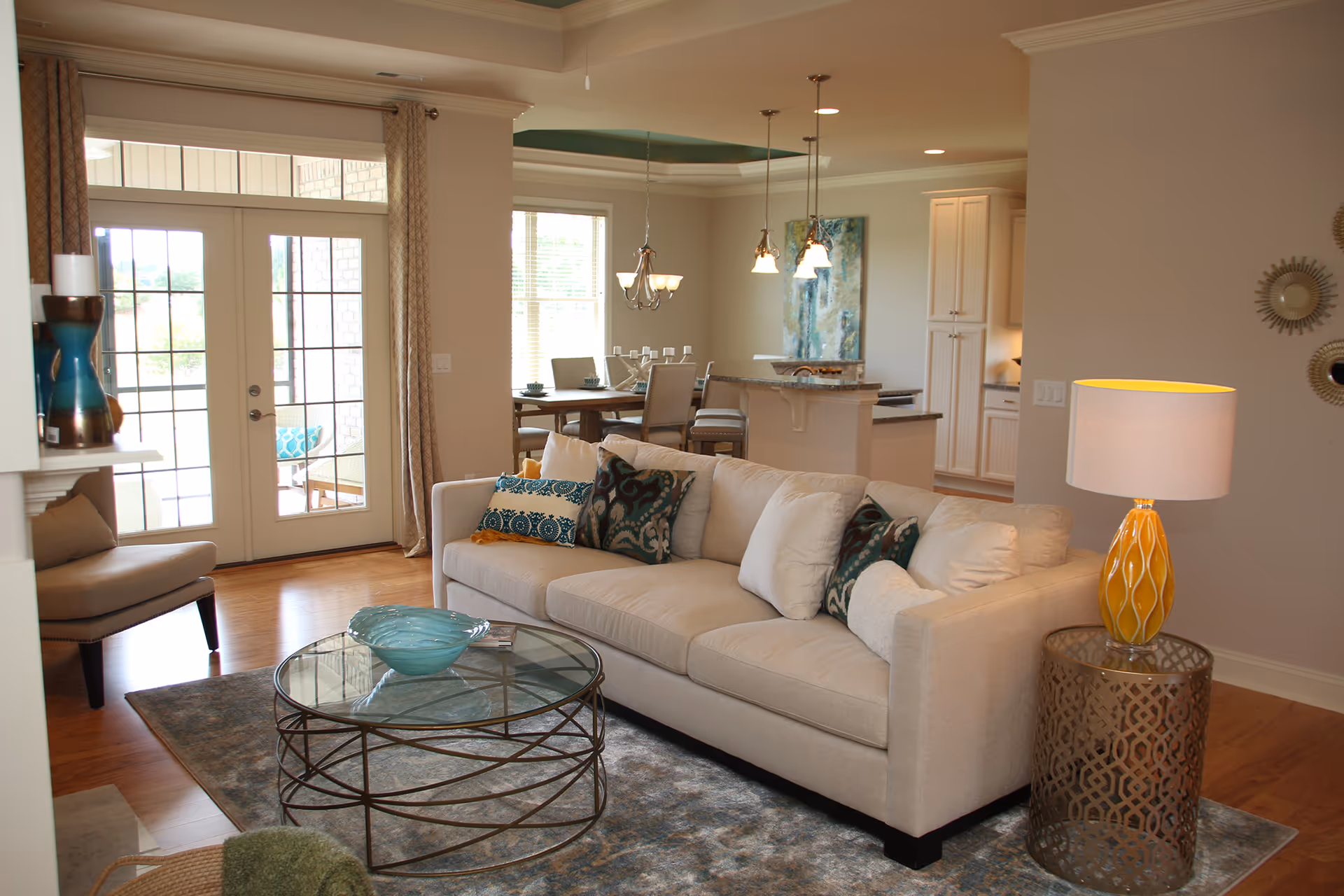 A cozy living room with a beige sofa adorned with patterned cushions, a round glass coffee table with a decorative bowl, a side table with a yellow lamp, and a chair near glass double doors leading to an outdoor area. In the background, there is a dining area with a table and chairs, pendant lights, and a kitchen with white cabinetry.