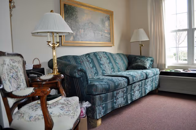 A patterned green sofa and upholstered chairs in a cozy living room with lamps, framed art, and a window letting in daylight.