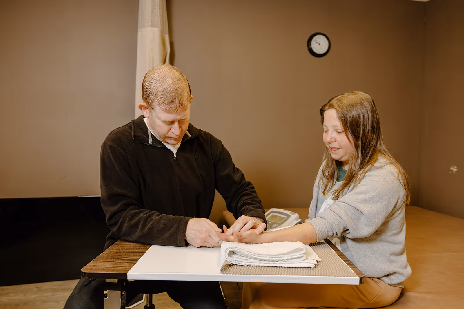 A healthcare professional is examining the hand of a woman seated at a table in a clinical room with brown walls. A clock is visible on the wall behind them, and a folded towel is on the table.