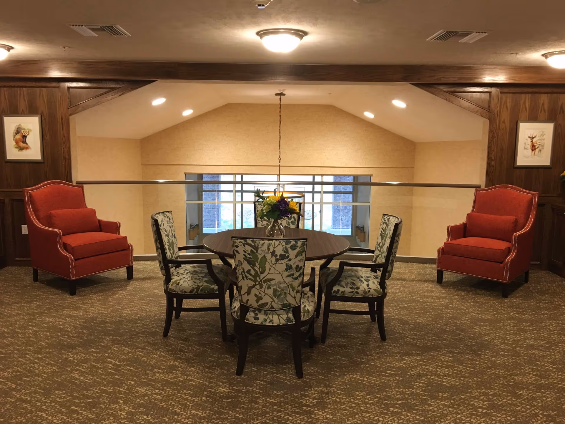 A cozy seating area in a retirement facility with a round wooden table surrounded by four floral-patterned chairs. Two red upholstered armchairs are placed against the wood-paneled walls on either side. The space is carpeted and softly lit with ceiling lights. There are framed pictures on the walls and a glass railing overlooking a lower level.