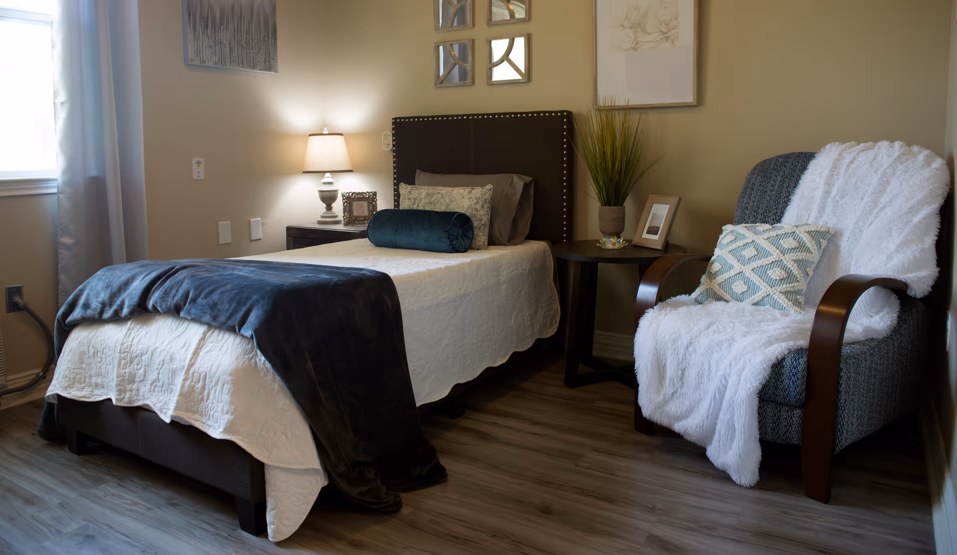 A cozy bedroom in a senior living facility featuring a single bed with white bedding and a dark blue throw blanket. Next to the bed is a nightstand with a lamp and a small framed picture. A comfortable armchair with a white throw blanket and a decorative pillow sits beside a round side table holding a potted plant and a framed photo. The room has light-colored walls, wooden flooring, and a window with curtains allowing natural light to enter.