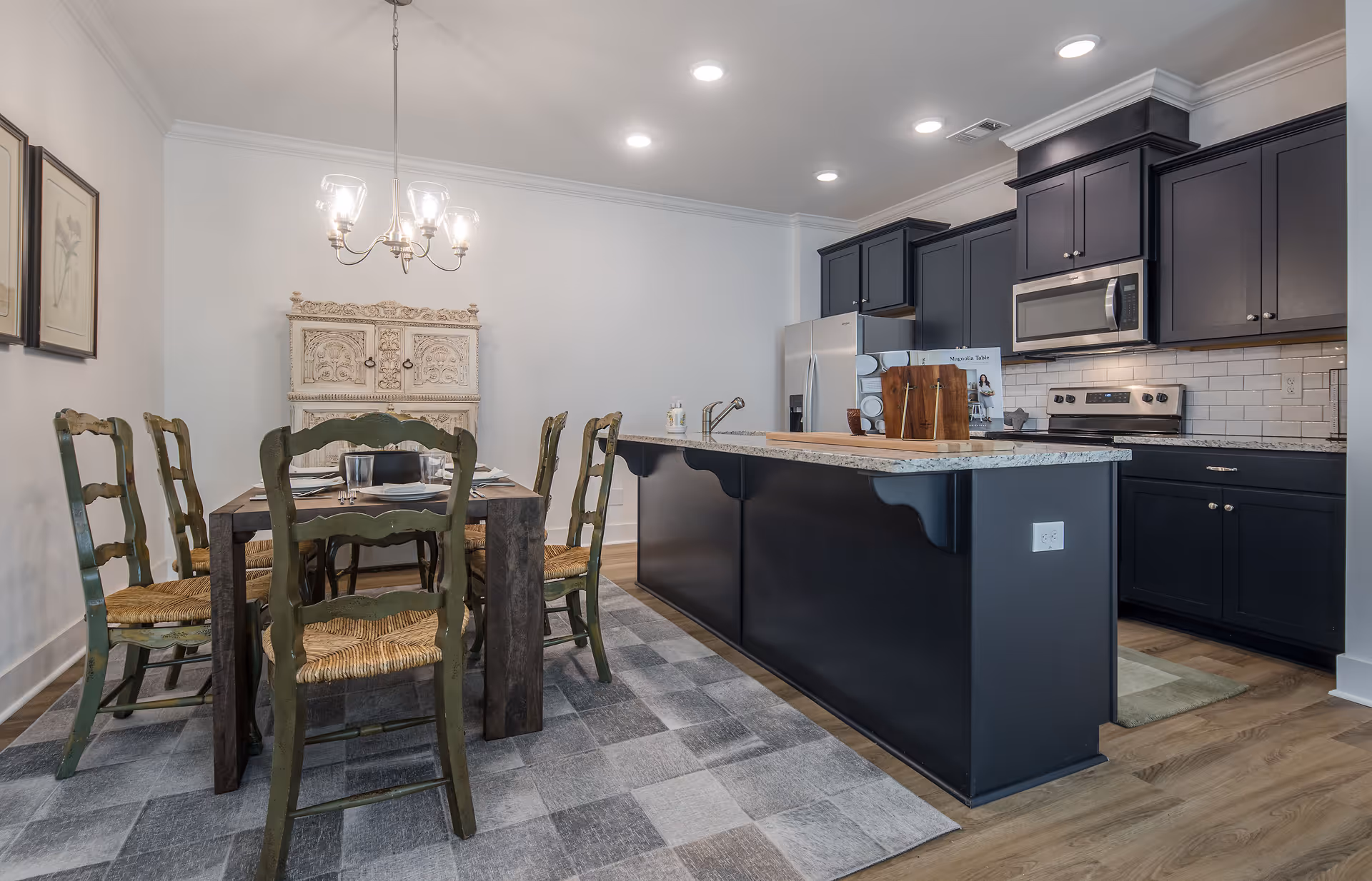 Open-plan dining area and kitchen featuring a wooden table with green chairs under a chandelier beside a dark island and matching cabinets.