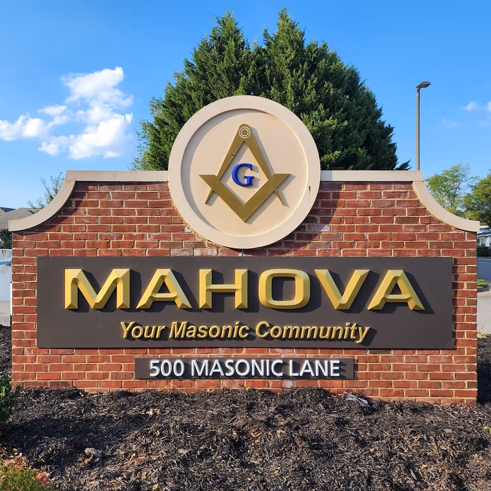 Brick sign for MAHOVA, Your Masonic Community, located at 500 Masonic Lane, with a Masonic square and compass symbol above the text, set against a backdrop of green trees and a blue sky.