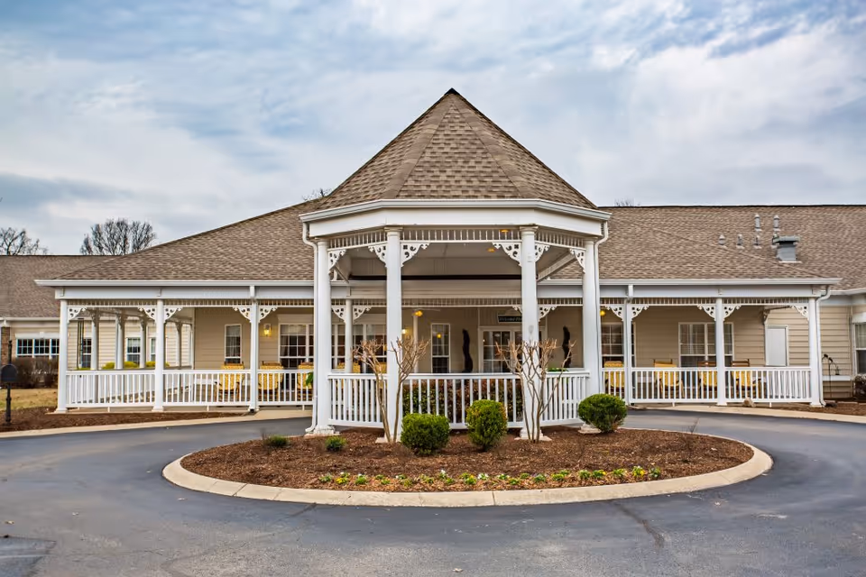 Front exterior view of a senior living facility with a covered entrance featuring white columns and decorative trim. There is a circular driveway with landscaped bushes and flowers in the center. The building has beige siding and a brown shingled roof under a cloudy sky.