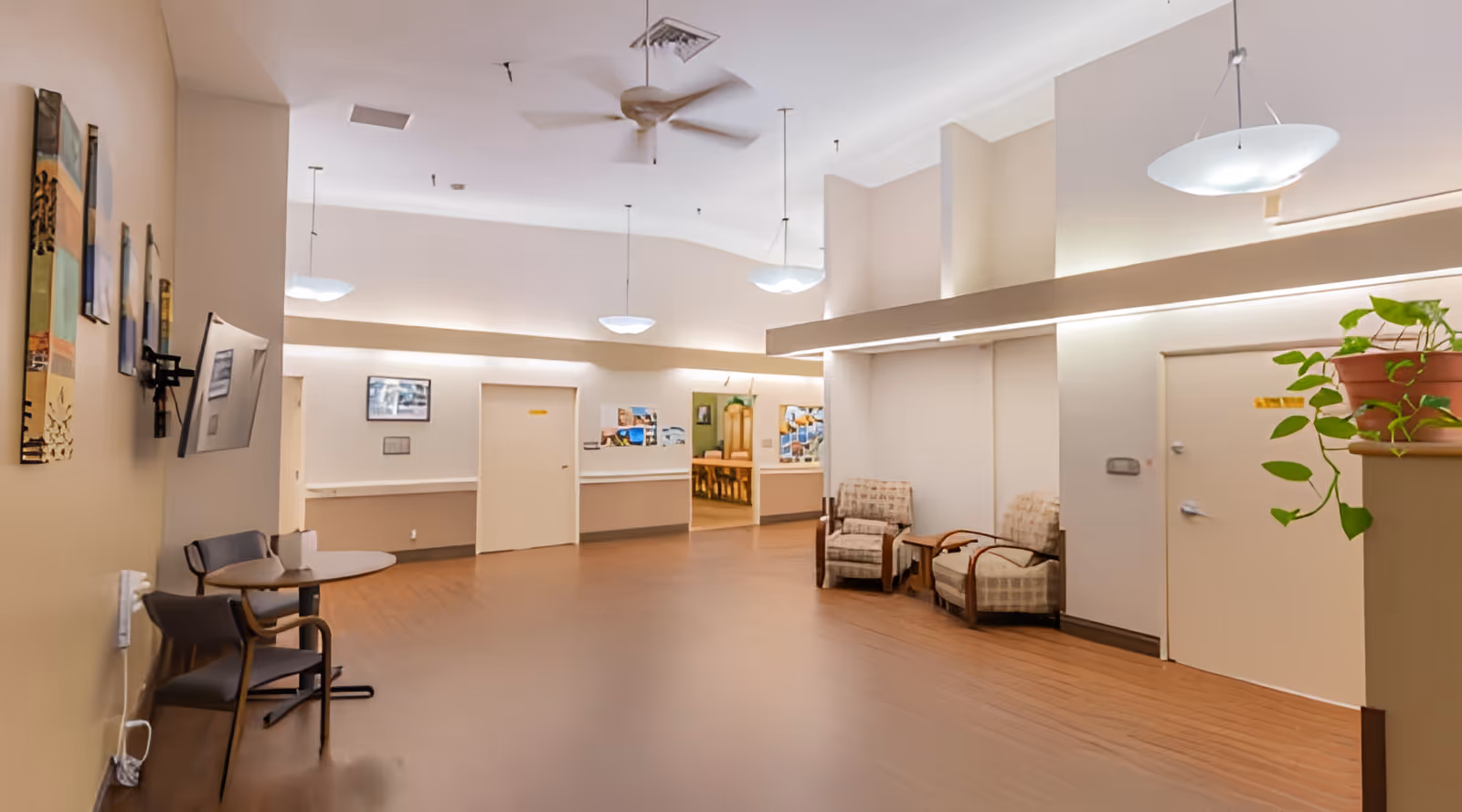 Well-lit senior living common area with chairs, tables, pendant lights, and a reception hallway.