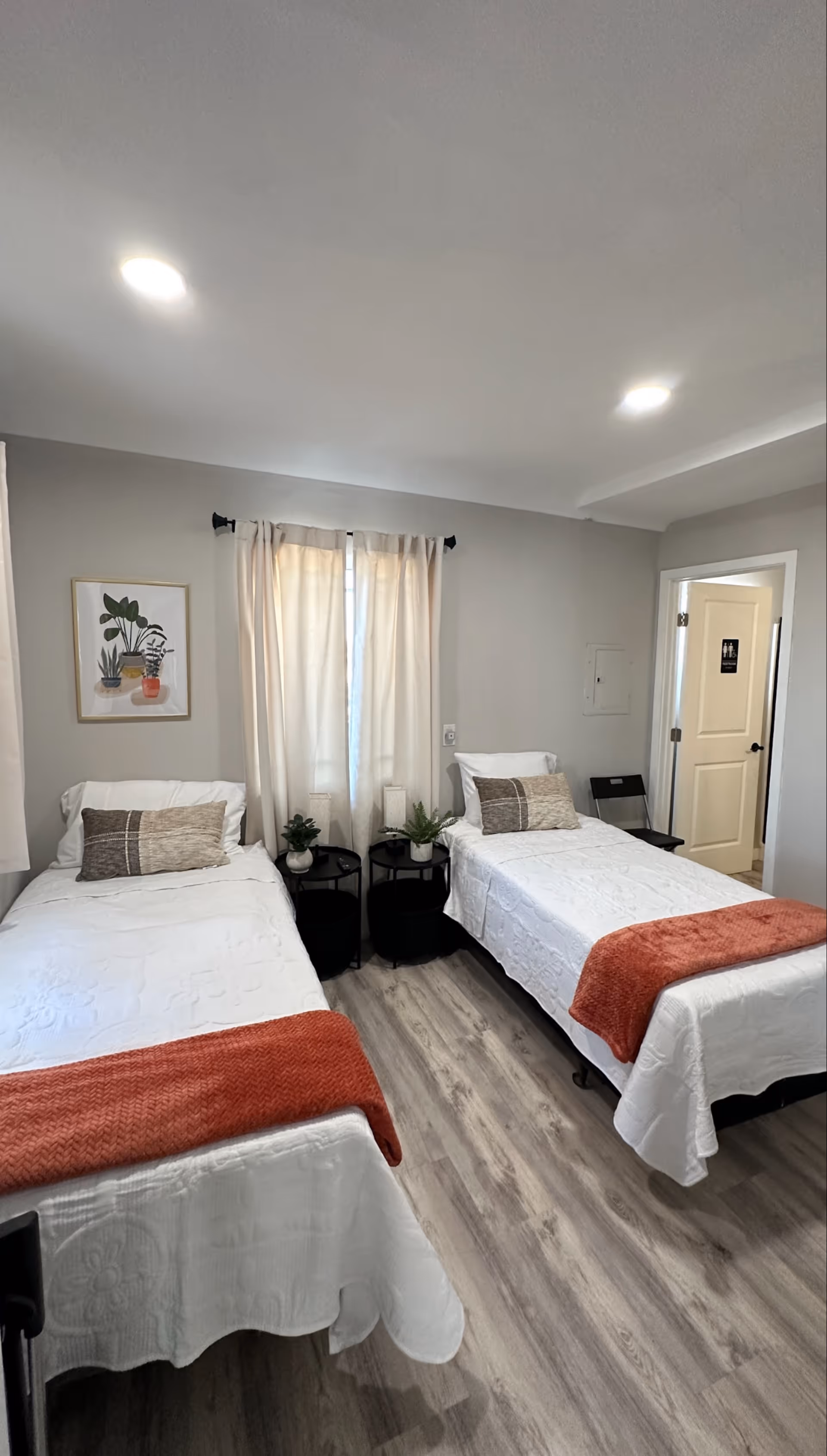 A bright and clean bedroom with two single beds, each with white bedding and a rust-colored throw blanket at the foot. Between the beds are two small black round tables with potted plants and a candle. A window with light beige curtains is centered on the wall behind the beds, and a framed botanical print hangs to the left of the window. The floor is a light wood laminate, and there is a door to the right leading to another room.