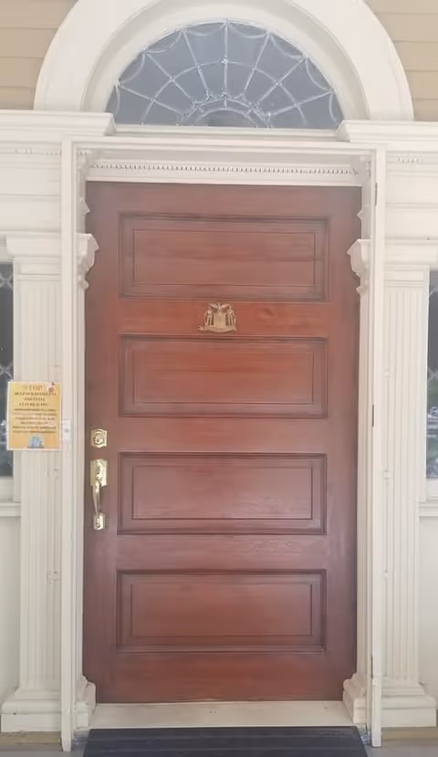 A large, solid wooden front door with decorative panels and a brass door knocker, framed by white columns and molding. Above the door is a semi-circular window with a decorative spiderweb-like design. A yellow sign is posted on the left side of the door frame.