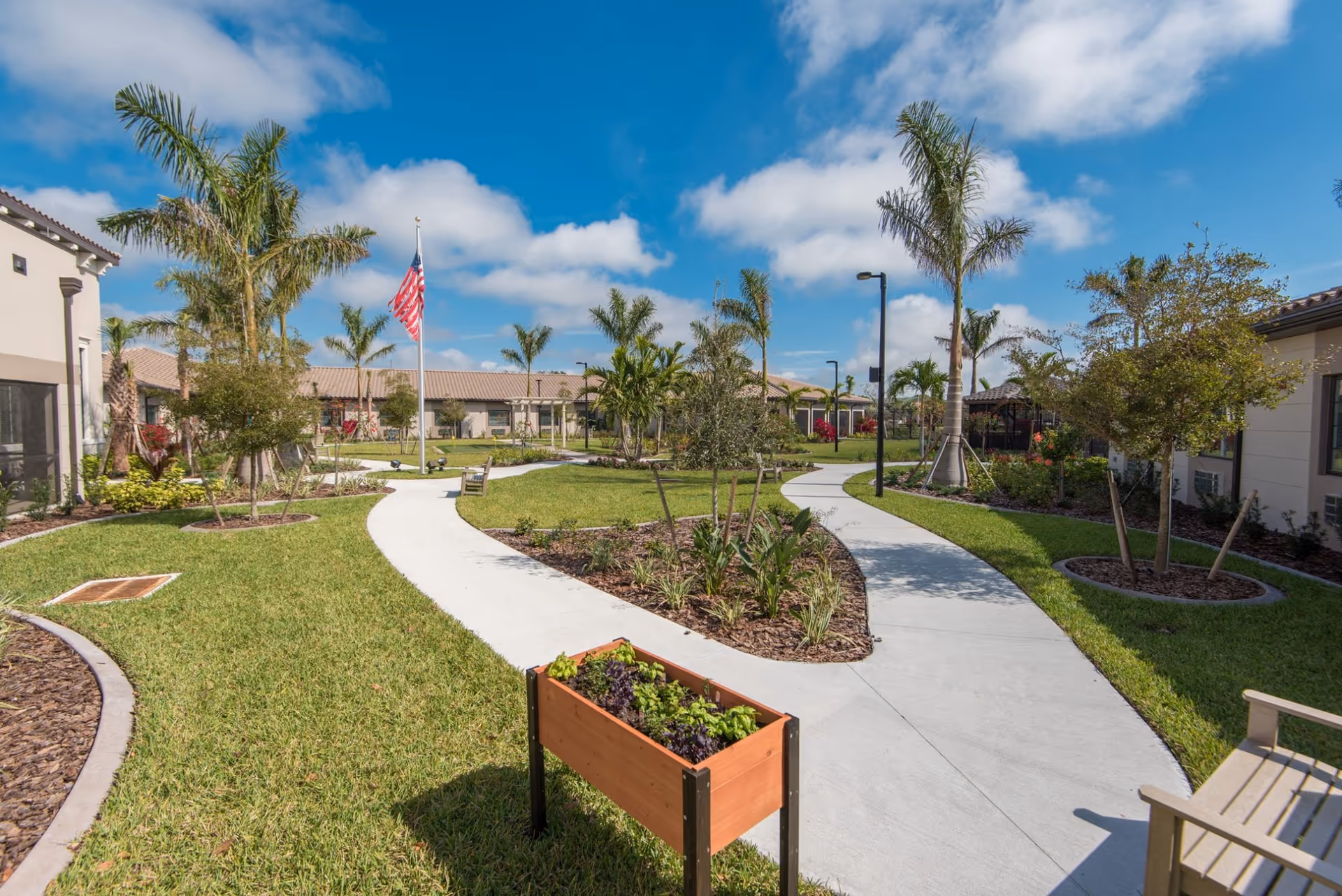 Outdoor garden area at Inspired Living Bonita Springs with paved walkways, green grass, palm trees, small plants, an American flag on a flagpole, and benches under a partly cloudy blue sky.