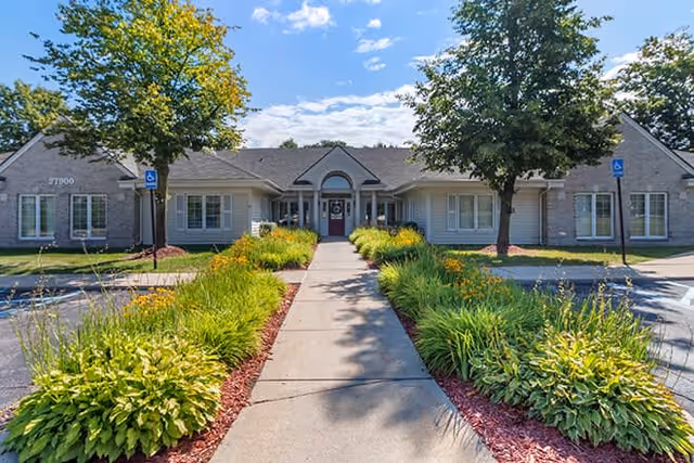 Front exterior view of a single-story senior living facility building with a paved walkway leading to the entrance. The building is surrounded by well-maintained landscaping with green bushes and trees under a partly cloudy blue sky.